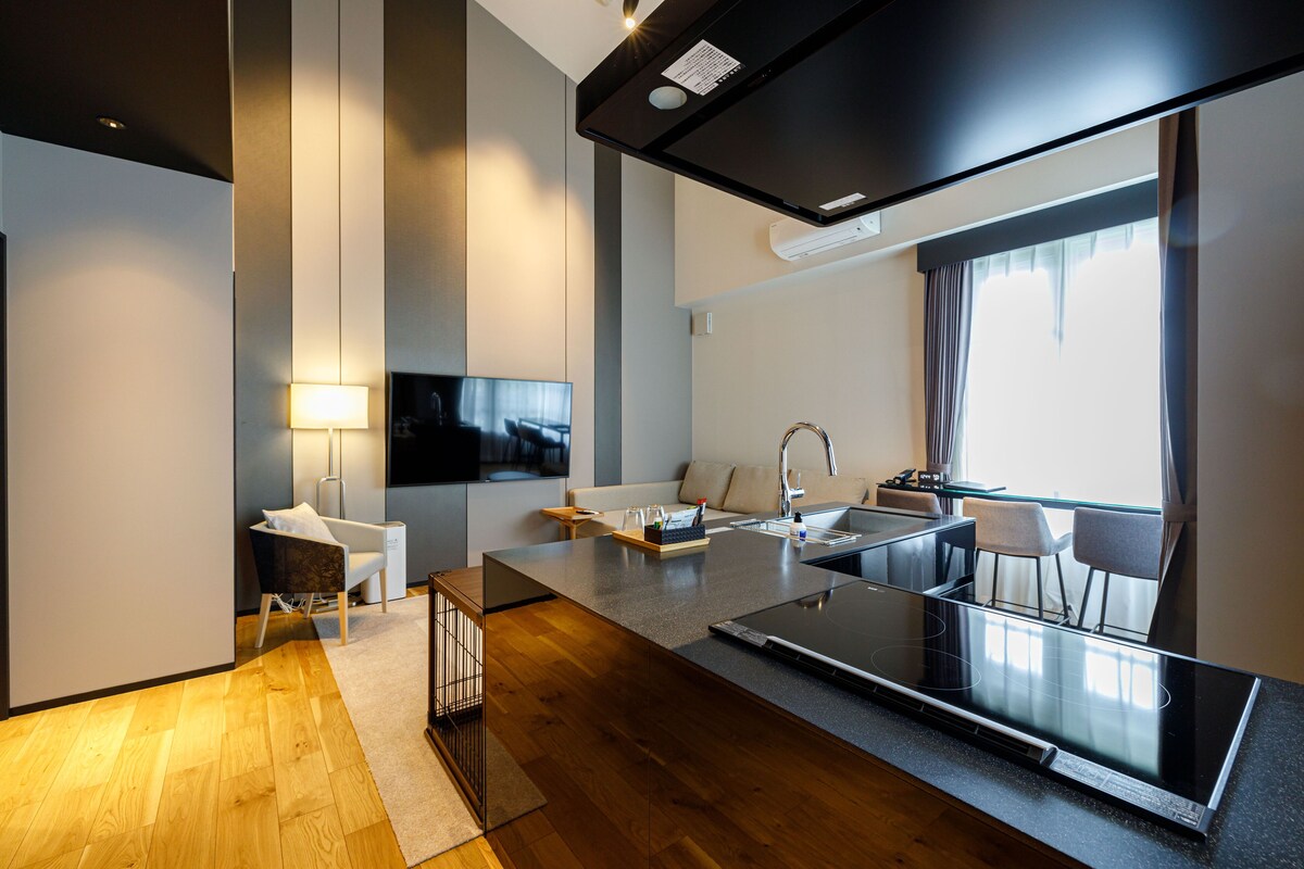 A modern kitchen with a large island, a built-in oven, and a sink. There is a flat-screen TV mounted on the wall, a sofa, and a dining area with chairs.