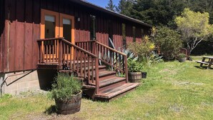 A rustic wooden building with a wooden staircase leading to a door, surrounded by greenery and trees.