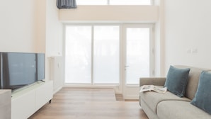 Ein modernes Wohnzimmer mit Flachbildfernseher, einer Couch mit Kissen und einer großen Schiebeglastür, die auf einen Balkon führt.