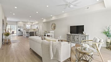 A spacious living room with a white sofa, a television, and a dining area in the background.