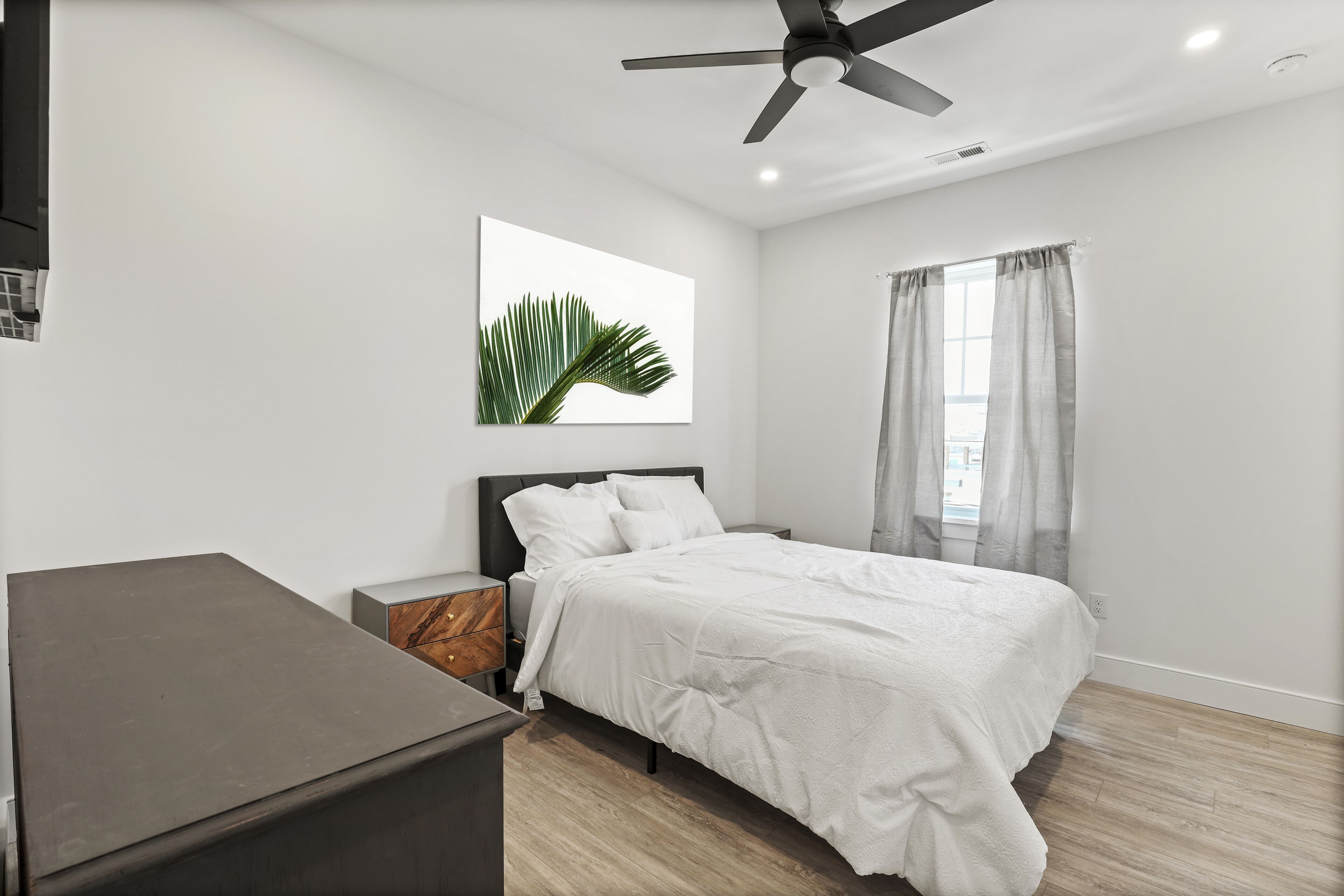 A bedroom with a bed, a nightstand, a desk, a ceiling fan, and a window with curtains.