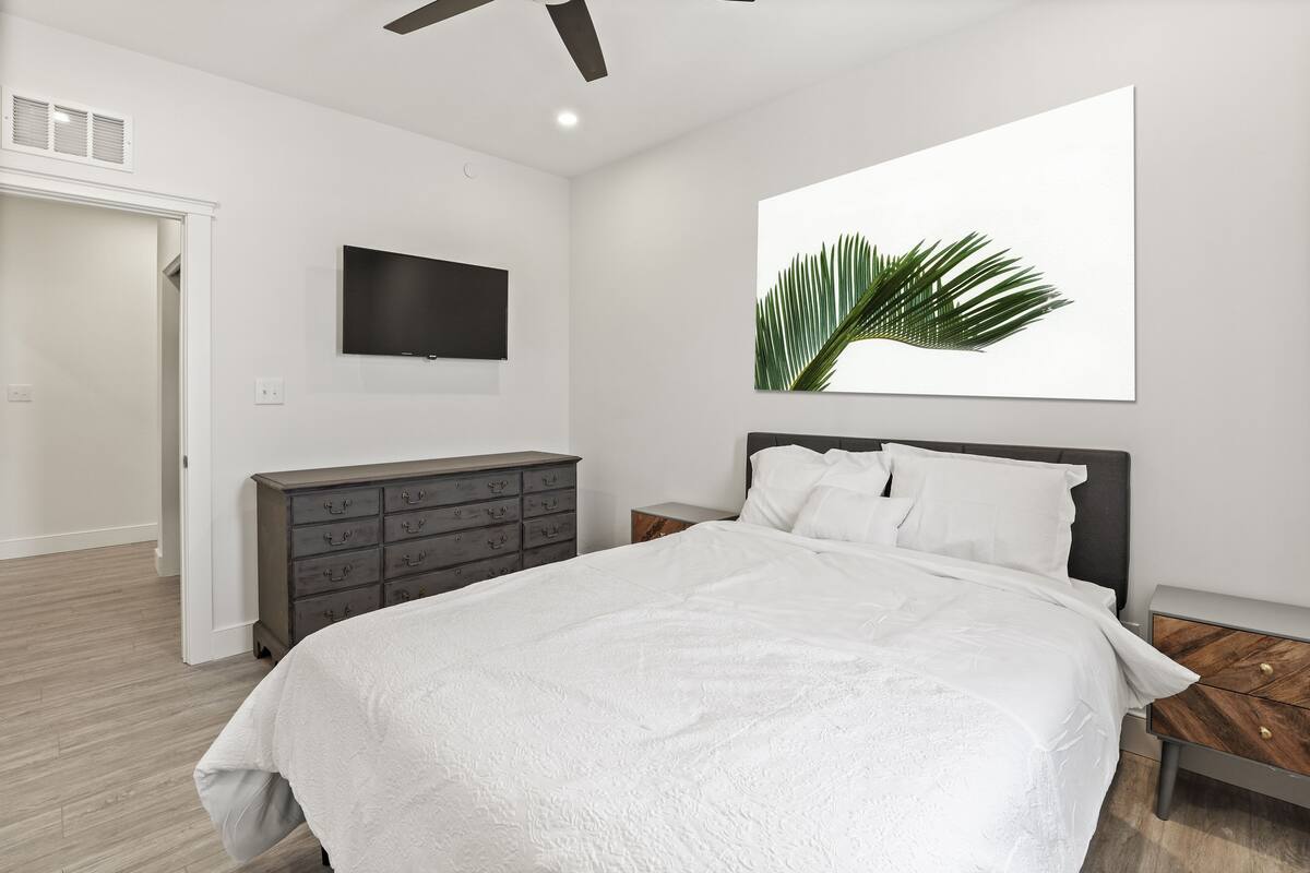A modern bedroom with a large bed, a flat-screen TV mounted on the wall, a wooden nightstand, and a large potted plant.