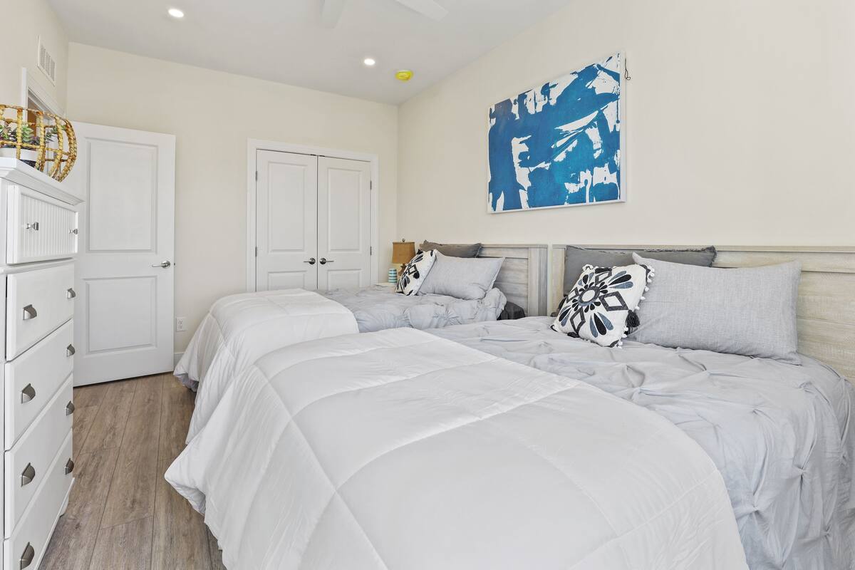 A bedroom with two beds, white bedding, a large abstract painting, and a wooden dresser.