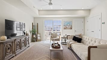 A spacious living room with a large flat-screen TV, a beige sofa, a white armchair, and a decorative rug.