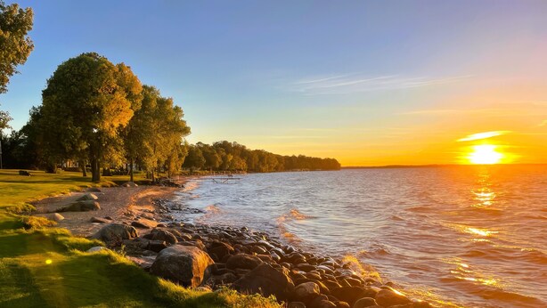 Een vredig uitzicht op het meer met een gazon, bomen en een heldere lucht tijdens zonsondergang.