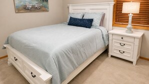 A bedroom with a bed, a nightstand, a lamp, a painting, and a window with blinds. - Lakeside Retreat at Izatys - Unit #230 – The Perfect Getaway for All Seasons! (Onamia)