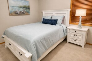 Een slaapkamer met een bed, nachtkastje, lamp, schilderij en een raam met jaloezieën.
