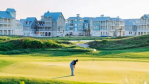 Ein Golfplatz, auf dem eine Person in blauem Hemd und weißen Hosen zum Putten ansetzt.