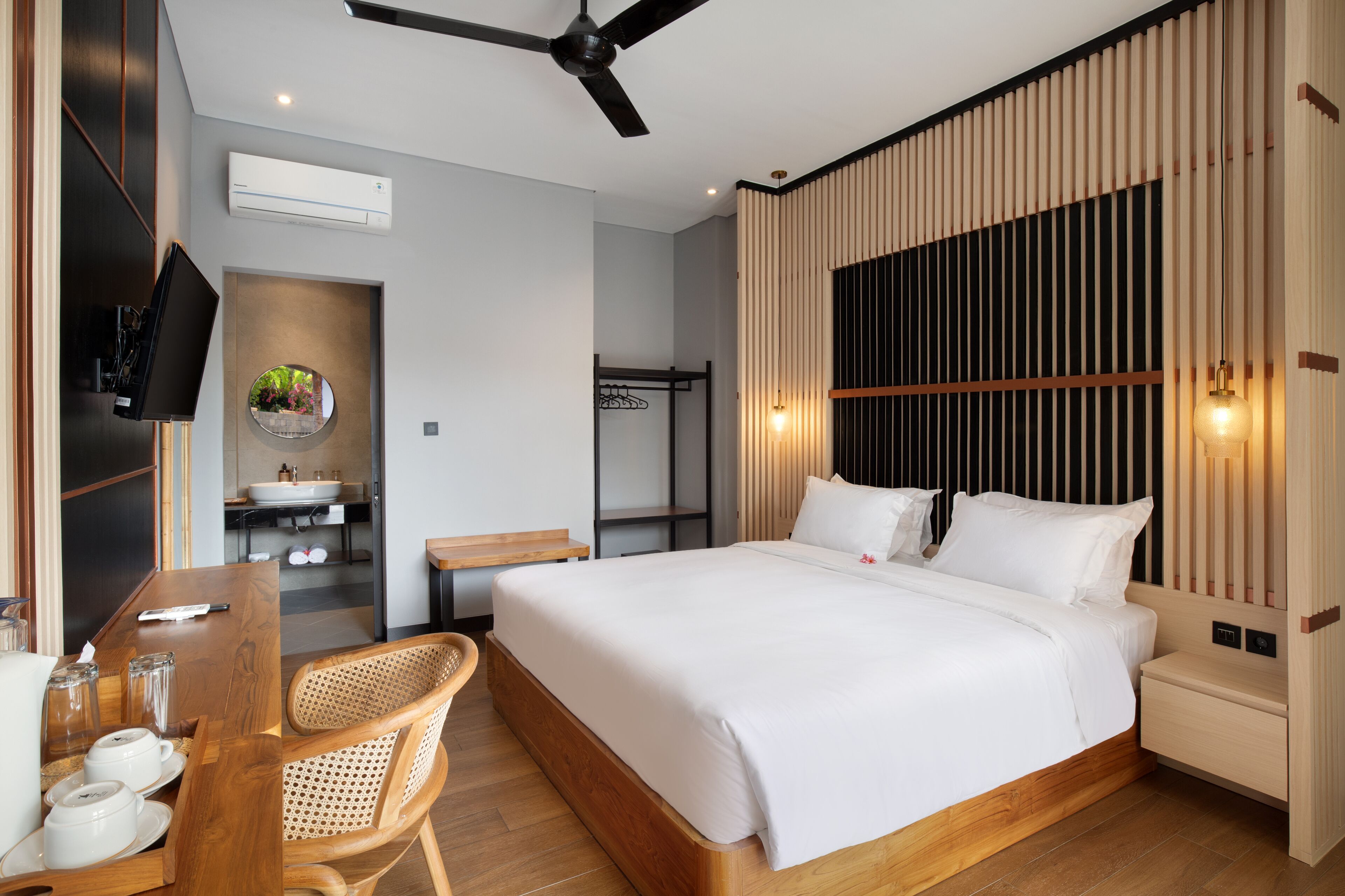 A modern hotel room with a large bed, a desk with a chair, a television, a bathroom with a sink, and a ceiling fan.