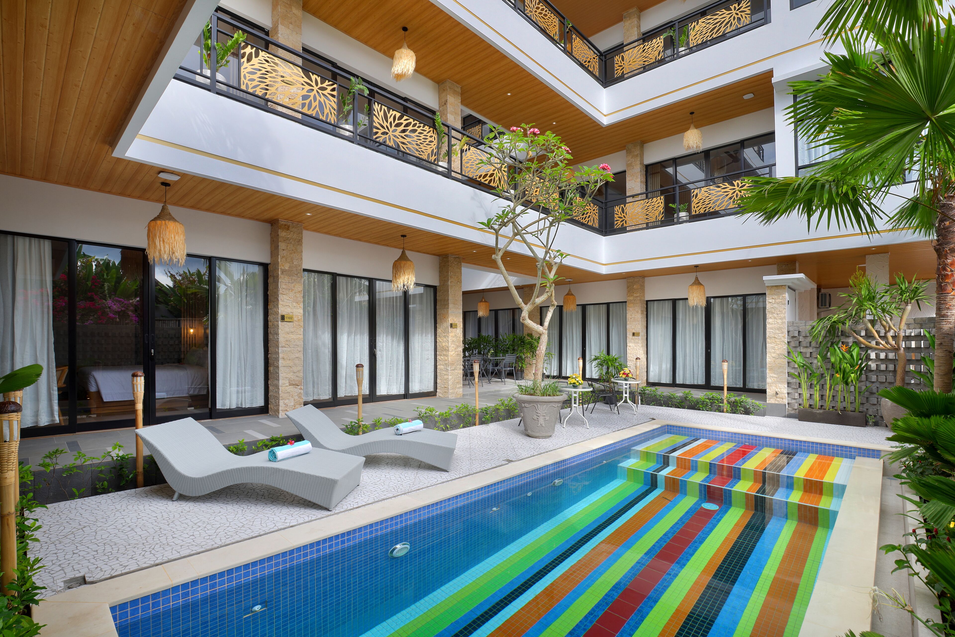 Un área de piscina interior moderna con un suelo de baldosas de colores, sillas de descanso y un árbol en el centro.