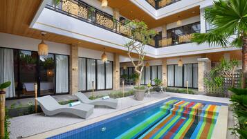 Un área de piscina interior moderna con un suelo de baldosas de colores, sillas de descanso y un árbol en el centro.