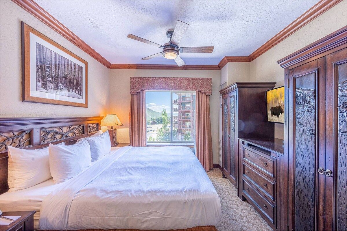 Quarto de hotel com cama, ventilador de teto, uma grande janela com cortinas e um rack para televisão.