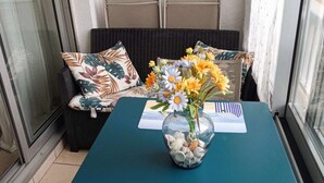 A balcony with a table set with a placemat, a vase with flowers, and a wicker sofa with cushions. - Studio in Pin Rolland, beach and shops on foot (ST MANDRIER SUR MER)
