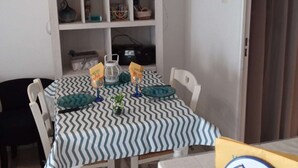 A dining area with a striped tablecloth, white chairs, a white cabinet with shelves, and a door leading to another room. - Studio in Pin Rolland, beach and shops on foot (ST MANDRIER SUR MER)