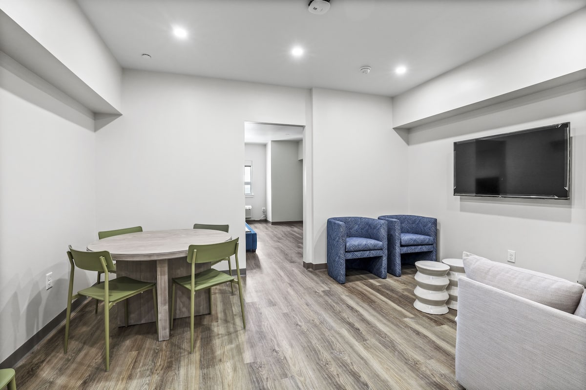 A modern, well-lit room with a wooden dining table, green chairs, a blue sofa, and a TV mounted on the wall.