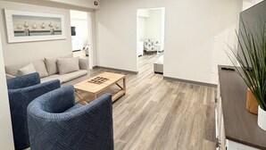 Un salon moderne comprenant un canapé, deux fauteuils, une table basse et un coin cuisine visible à travers une porte ouverte.