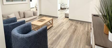 Una sala moderna con un sofá, dos sillones, una mesa de centro y una cocina visible a través de una puerta abierta.