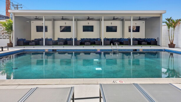 Un espace piscine moderne doté d’une piscine à eau claire, de chaises longues, de ventilateurs de plafond et d’une structure de bâtiment blanche.
