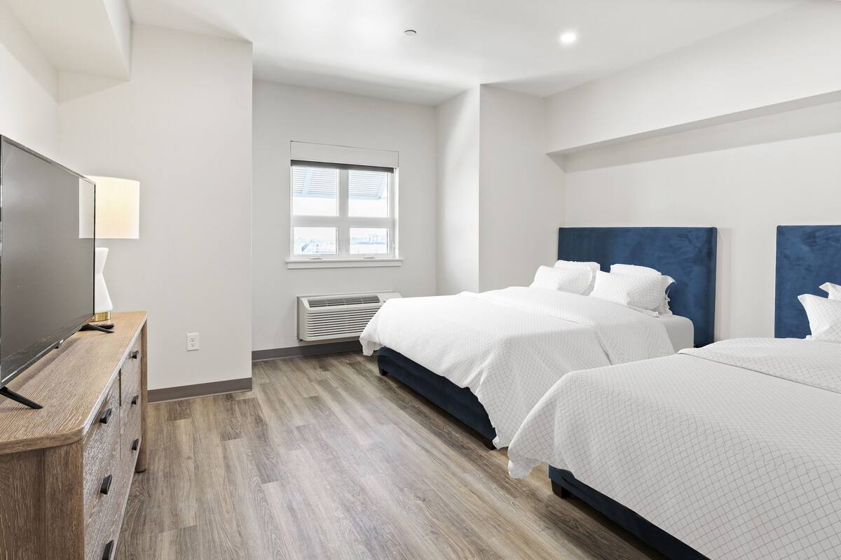 A hotel room with two beds, a wooden dresser, a flat-screen TV, and a window with blinds.