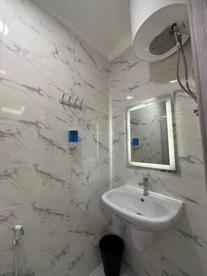 A bathroom with a marble wall, a sink, a mirror, a shower head, and a trash bin.