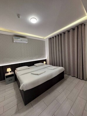 A modern hotel room with a large bed, bedside tables, an air conditioning unit, and a ceiling light.
