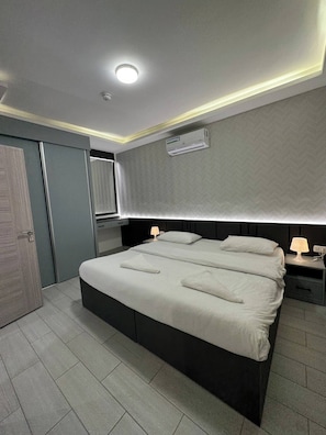 A modern bedroom with a large bed, bedside lamps, a ceiling light, and an air conditioning unit.