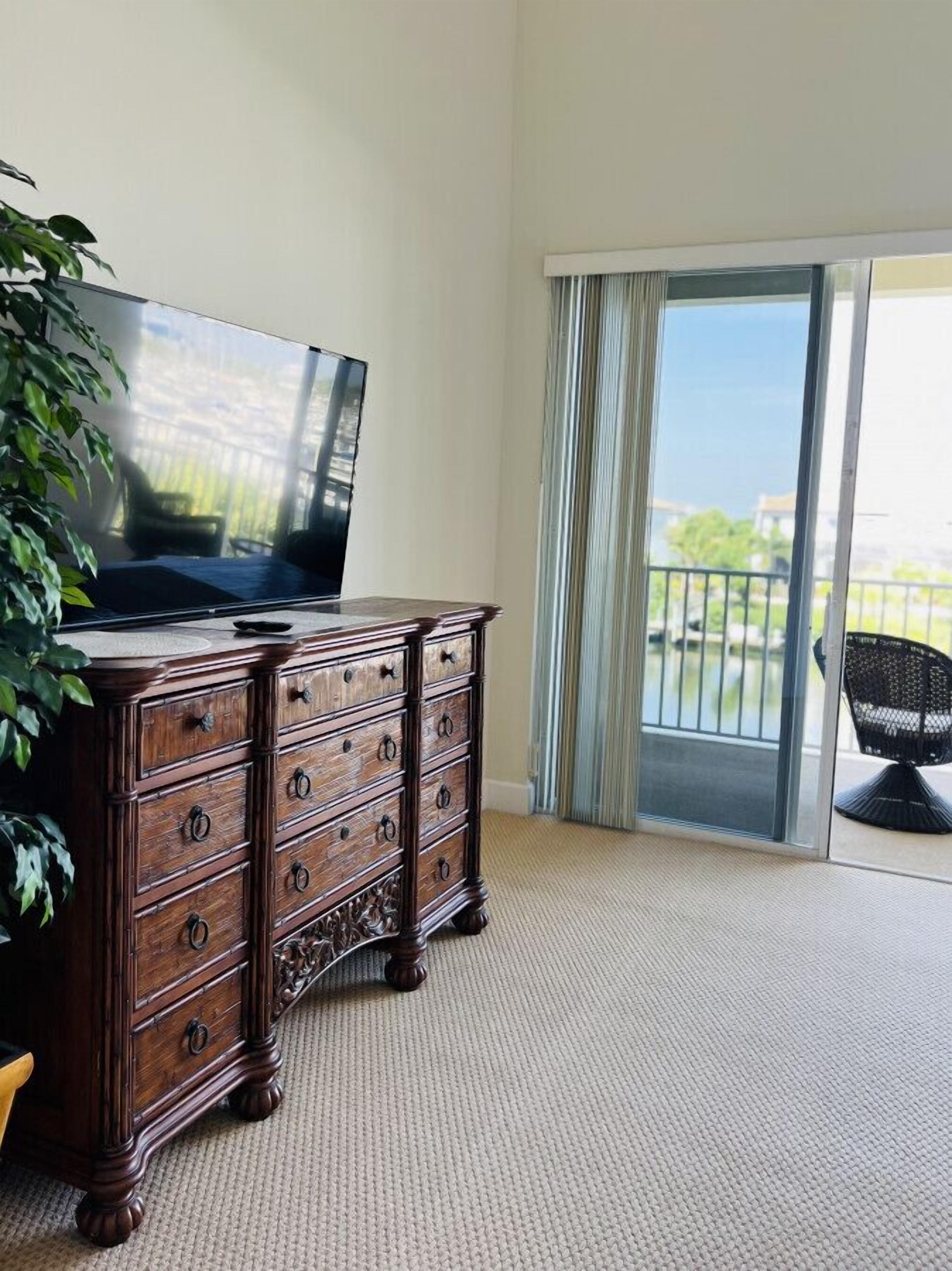 Quarto com cômoda de madeira, televisão em cima e vista para uma varanda com cadeira.