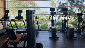 Ein Fitnessstudio mit Laufbändern, Crosstrainern und Kraftstationen, mit großen Fenstern, die einen Blick auf Palmen und Grünflächen bieten.