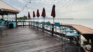Eine Holzterrasse mit Stuhl- und Tischenreihen, roten Sonnenschirmen und Blick auf das Wasser.