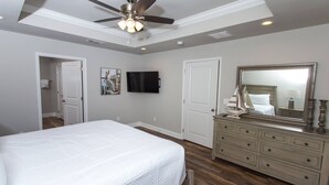 A bedroom with a bed, dresser, ceiling fan, TV, and a door leading to another room.