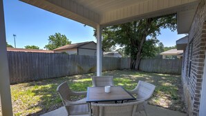 A covered patio with wicker chairs and a table, a wooden fence, and a brick wall in the background.