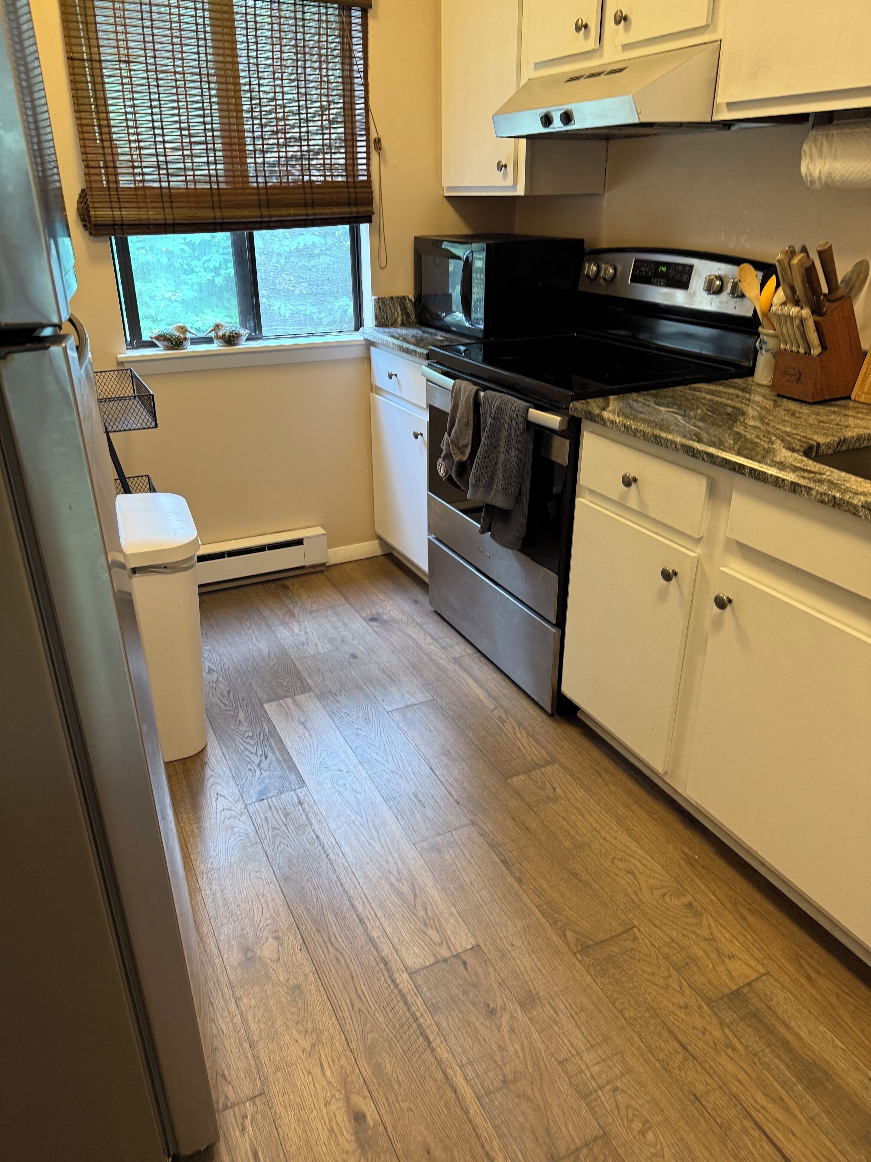 A kitchen with white cabinets, a stainless steel oven, a microwave, and a window with blinds.