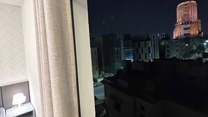 A hotel room with a large window, a bed, a bedside table with a lamp, and a radiator.