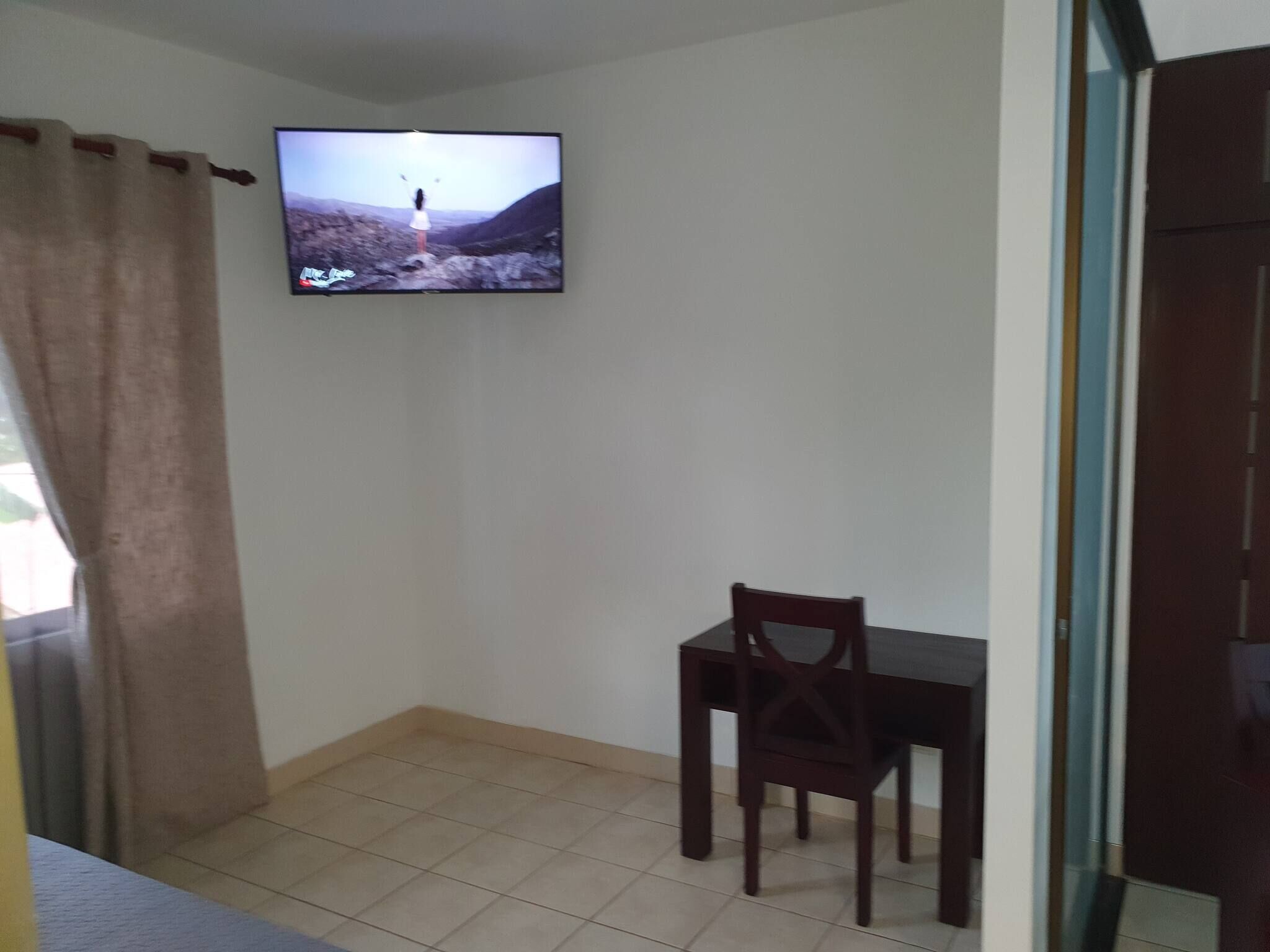 Une pièce avec un téléviseur fixé au mur, une table à manger avec une chaise et un rideau.