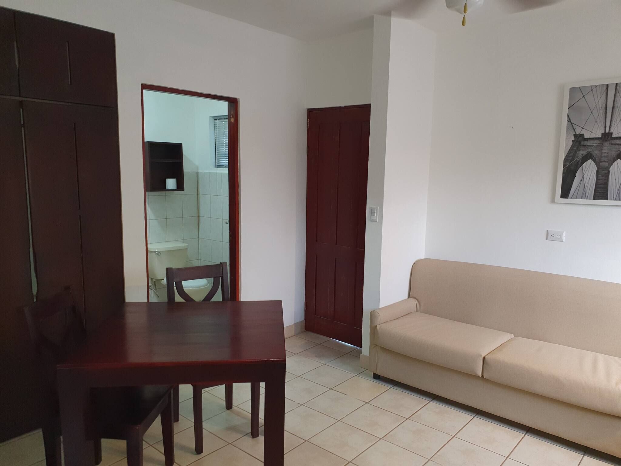 A living room with a wooden table and a beige sofa, a wooden wardrobe, and a door leading to a bathroom.