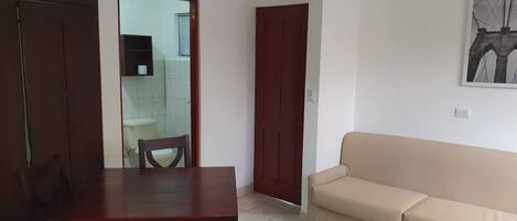 A living room with a wooden table and a beige sofa, a wooden wardrobe, and a door leading to a bathroom.