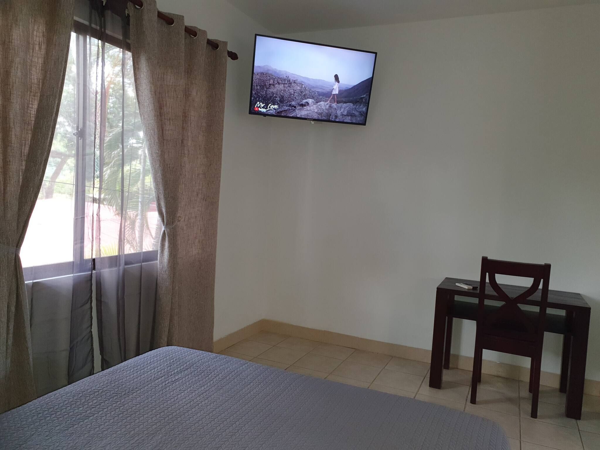 A room with a bed, a TV on the wall, a chair, and a window with curtains.