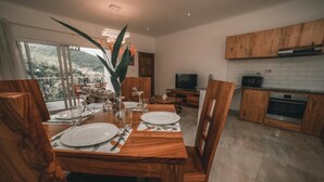A dining area with a wooden table set for four, a TV in the background, and a kitchen with wooden cabinets. - Kannel Apartments Seychelles - 2 Bedroom (Mahé Island)