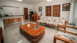 A modern living room with a wooden coffee table, a sofa with a tropical print, a dining area with wooden chairs, and a kitchen with a whiteboard. - Kannel Apartments Seychelles - 2 Bedroom (Mahé Island)