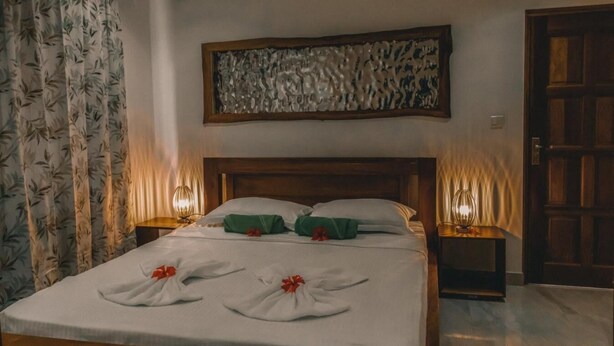 A neatly made bed with towels and a red bow, a wooden headboard, a wall art piece, and two bedside lamps. - Kannel Apartments Seychelles - 2 Bedroom (Mahé Island)