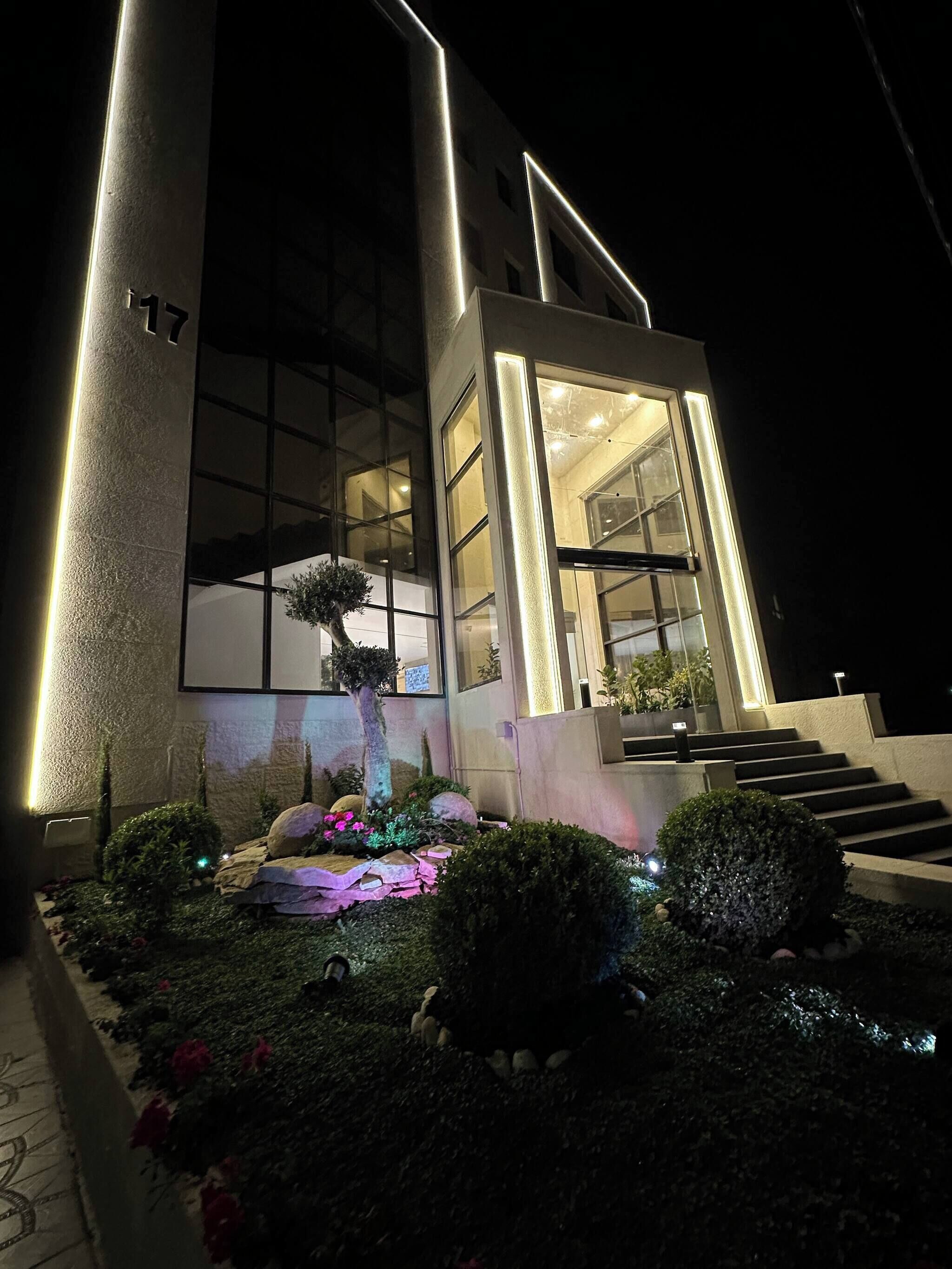 A modern building with a large glass facade, a well-lit entrance, and a landscaped garden with shrubs and a small tree.