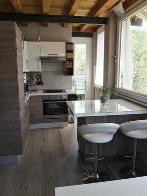 A modern kitchen with wooden cabinets, a built-in oven, a large island with bar stools, and a window providing a view of greenery. - Open Space in the Village With Direct Access to the Beach (Manerba del Garda)