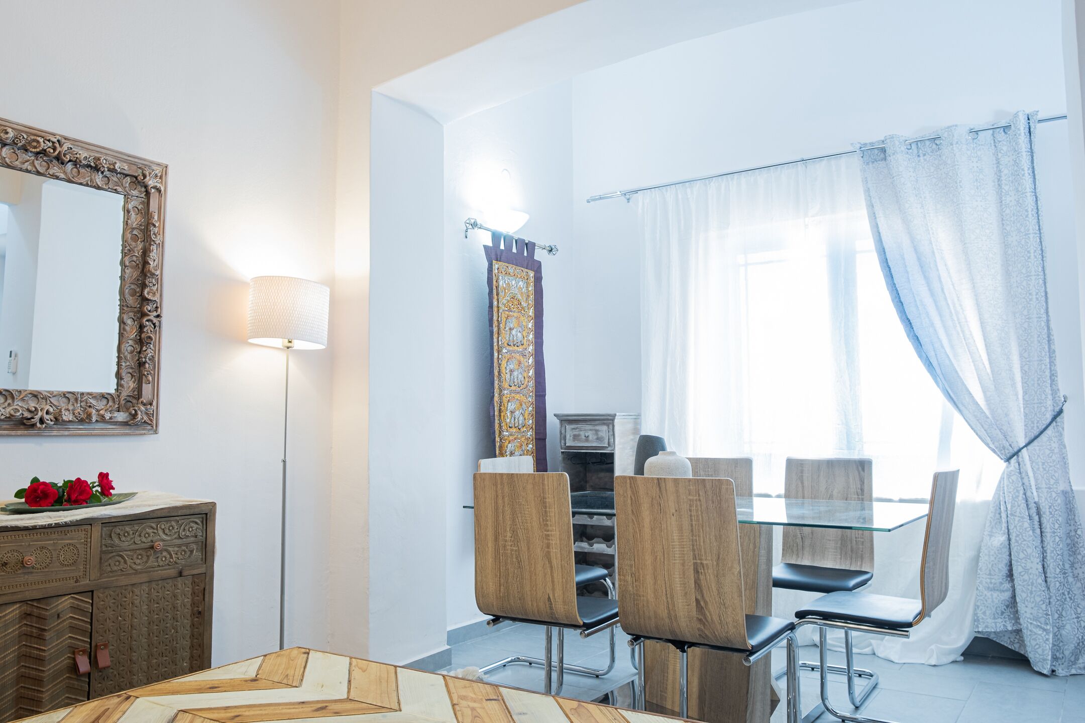 A modern dining area with a glass table, wooden chairs, and a decorative mirror on the wall.