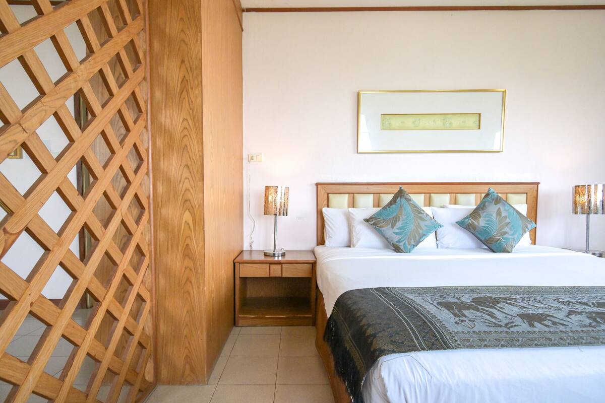 A hotel room with a wooden lattice partition, a bed with white bedding and decorative pillows, a bedside table with a lamp, and a framed picture on the wall.
