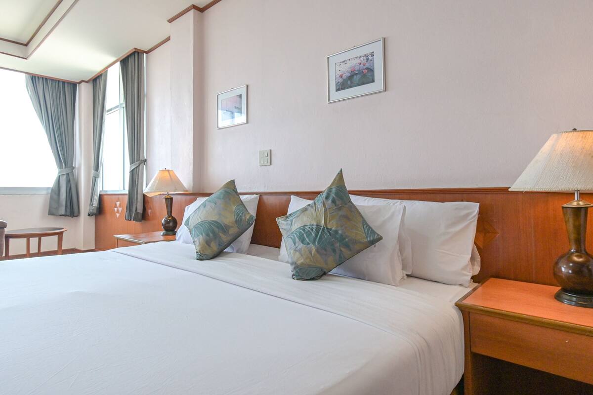 A hotel room with a large bed, two pillows, a nightstand with a lamp, a small table, and a window with curtains.
