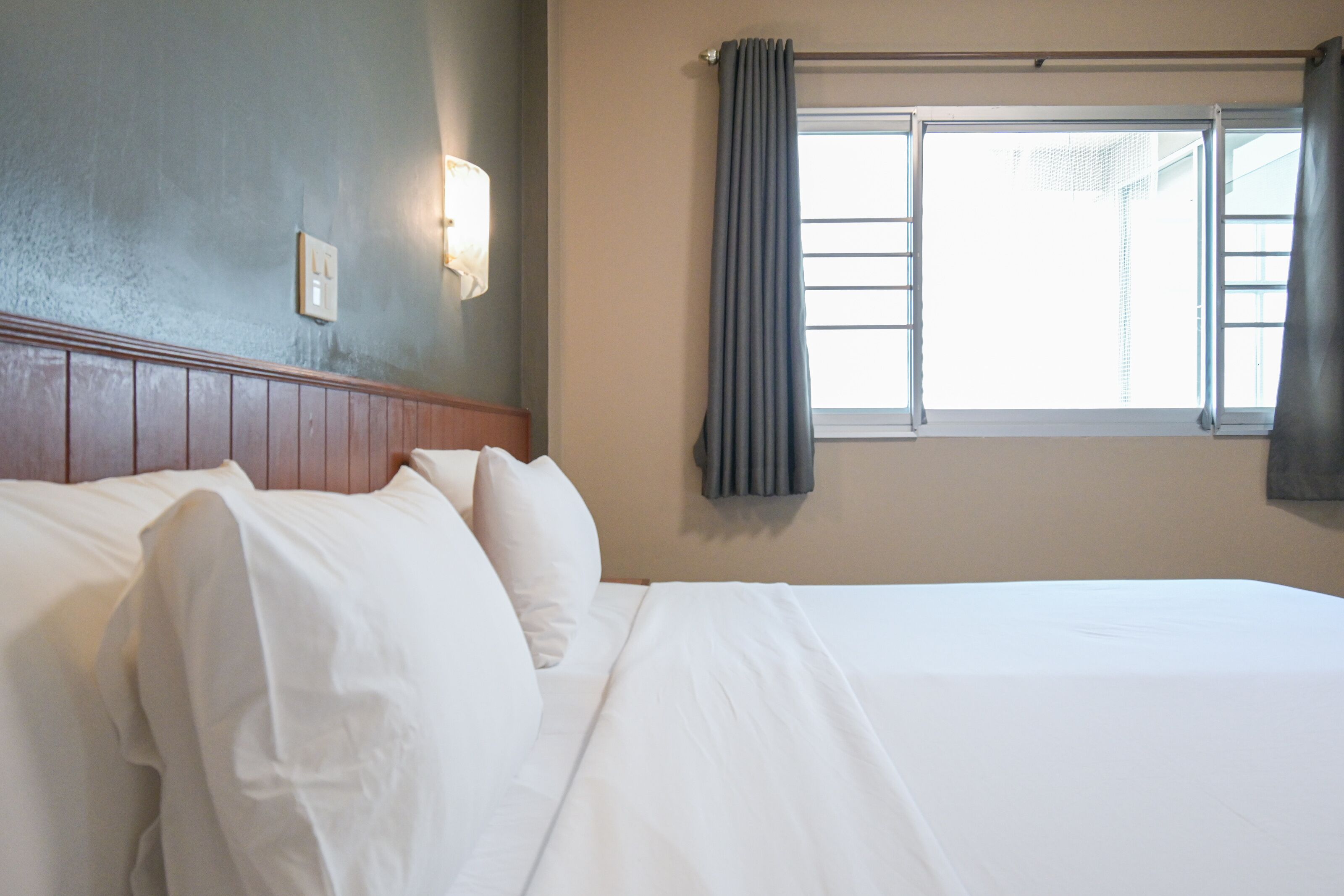 A hotel room with a bed, a headboard, a wall-mounted light, and a window with curtains.