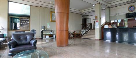 A spacious lobby with a leather sofa, a glass coffee table, a large wooden pillar, a reception desk, and a staircase.