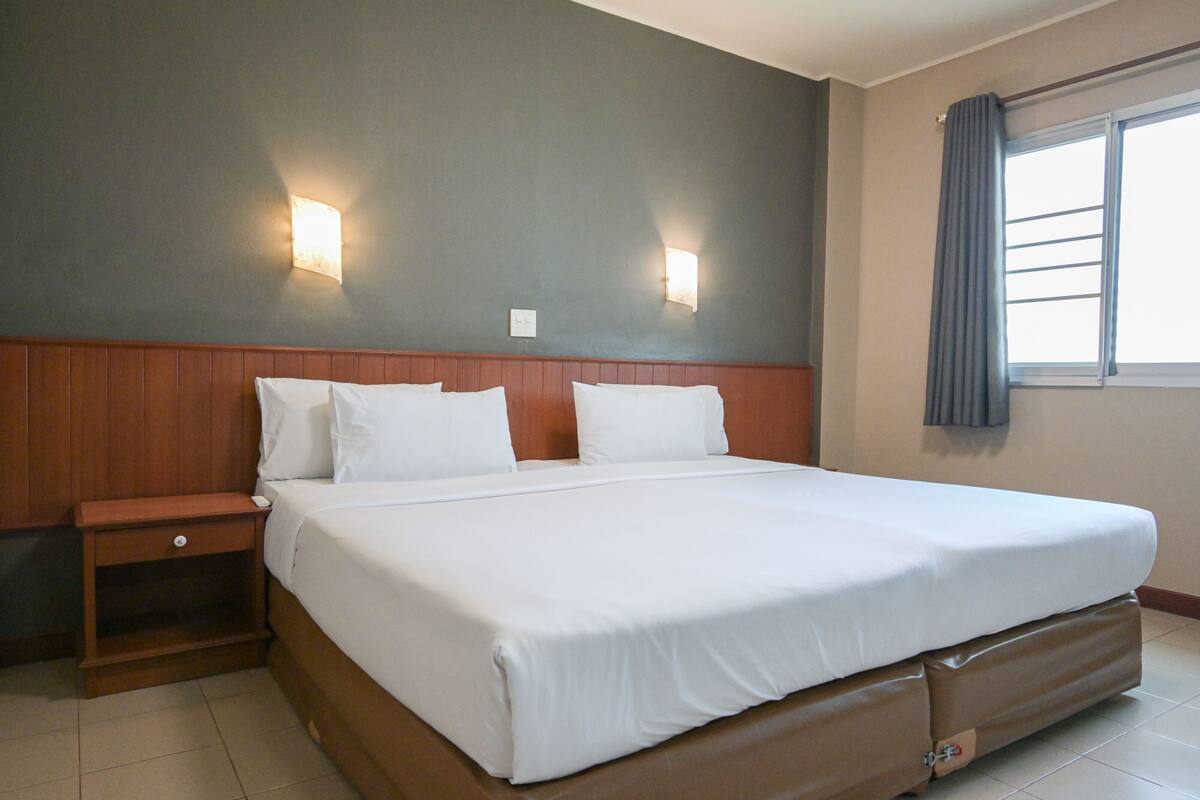 A hotel room with a large bed, wooden headboard, two wall-mounted lights, a nightstand, and a window with curtains.