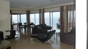 Amplia sala de estar con un gran ventanal corredero que da a un balcón con vistas al mar. Cuenta con un comedor con mesa y sillas, y un sofá en la sala.