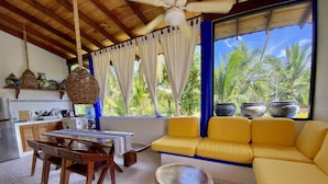 A cozy room with a wooden ceiling, a dining area with a table and chairs, a yellow sofa, and a window with a view of palm trees.